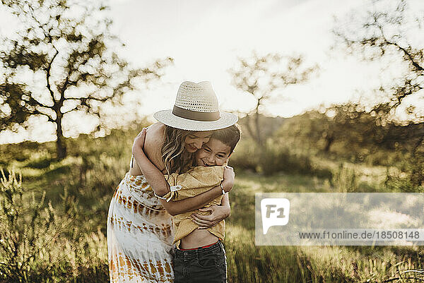 Youthful mother hugging son in sunny meadow with hat on