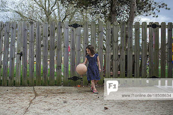 a small girl dribbles a basketball in a driveway in front of fence