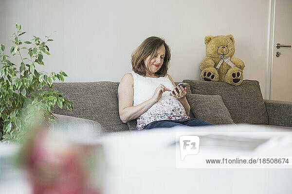 Pregnant woman sitting on sofa and using on mobile phone  Munich  Bavaria  Germany