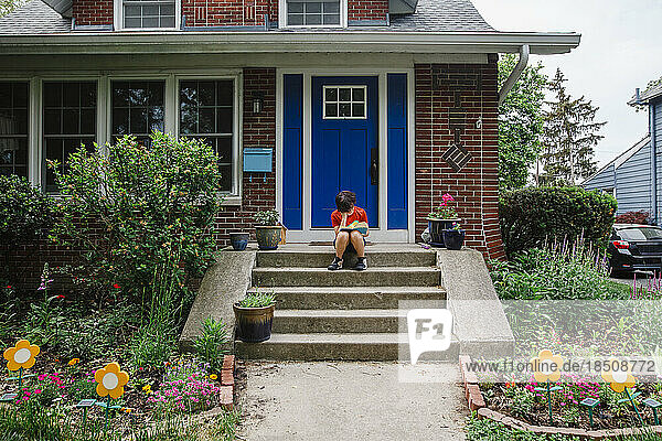 An unrecognizable child sits on stoop of home reading a book