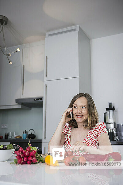 Pregnant woman talking on smartphone and sitting on chair with fresh vegetables in the kitchen  Munich  Bavaria  Germany