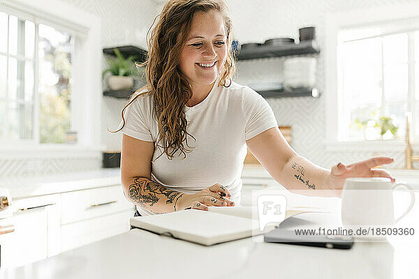 Smiling grown woman journaling and reaching for white mug of coffee