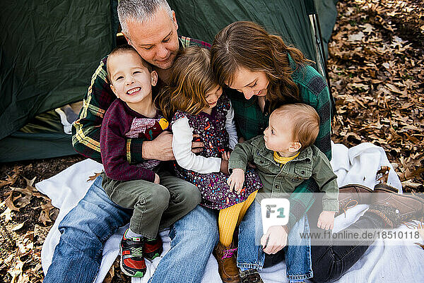 Smiling family sits on blanket outside of tent in fall leaves hugging