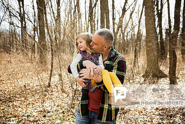 Loving father holds and kisses smiling daughter on cheek in Fall woods