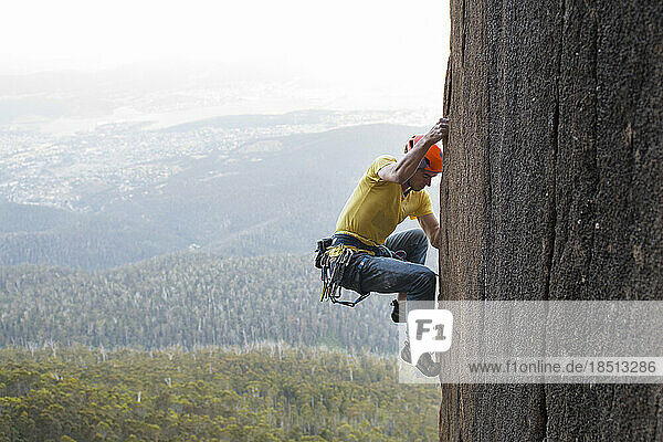 Side view of a male rock climber moving his feet up as he works his way up a diorite pillar in The Organ Pipes  at Mt Wellington  with the city of Hobart  Tasmania  seen in the bac