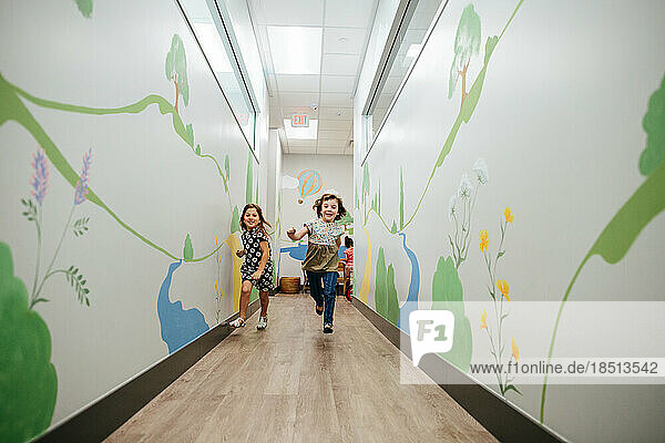 Two little girls smile and run down a mural covered hall in a school