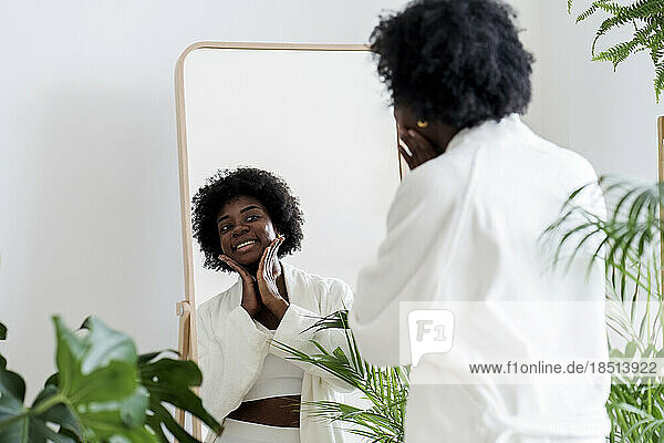 Smiling young woman applying moisturizer on face looking in mirror