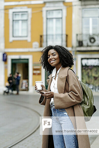 Happy young woman standing with mobile phone and coffee cup by tramway