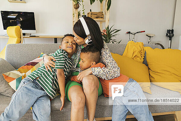 Mother wearing headphones embracing boys on sofa at home