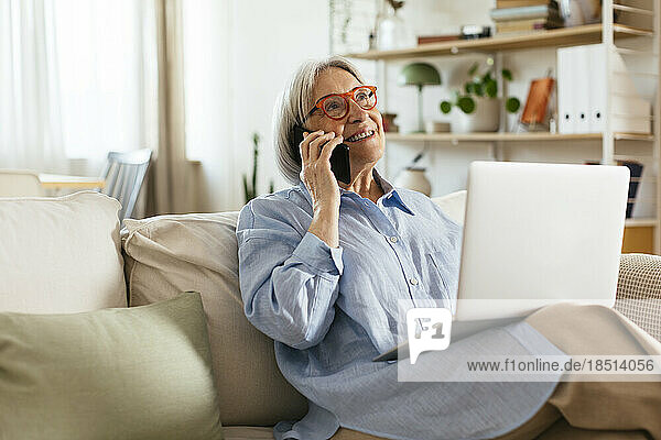 Smiling woman talking on smart phone sitting with laptop in living room