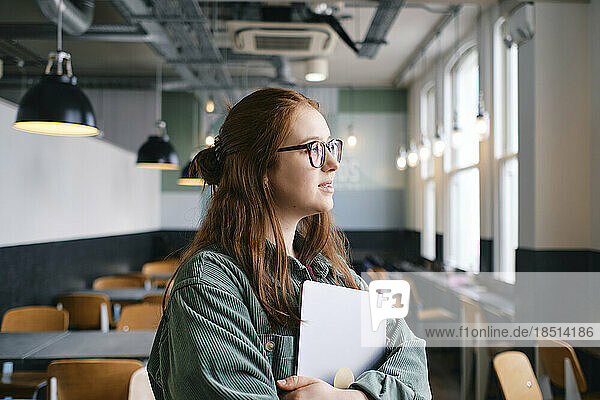 Thoughtful young freelancer standing with laptop in cafe