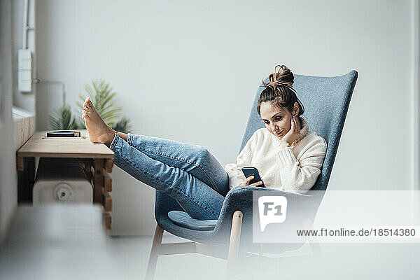 Young woman using smart phone sitting in armchair at home