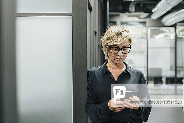 Blond businesswoman using mobile phone leaning on wall at office