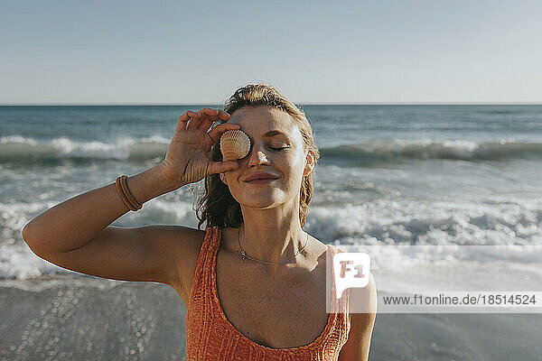 Smiling woman holding seashell over eye in front of sea