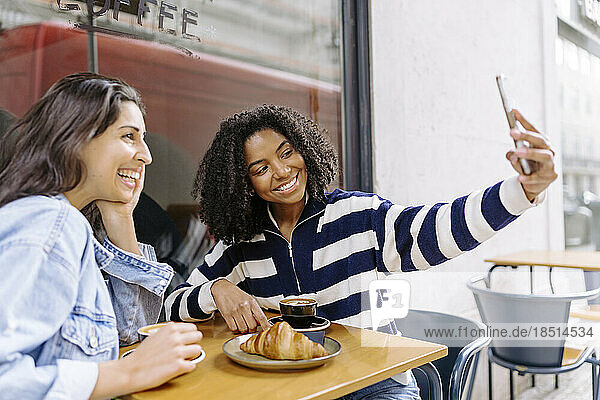 Smiling young woman taking selfie with friend at sidewalk cafe