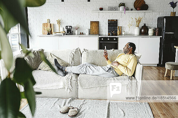 Mature man using smart phone lying on sofa at home