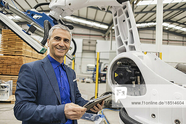 Smiling mature engineer standing with tablet PC by robotic arm