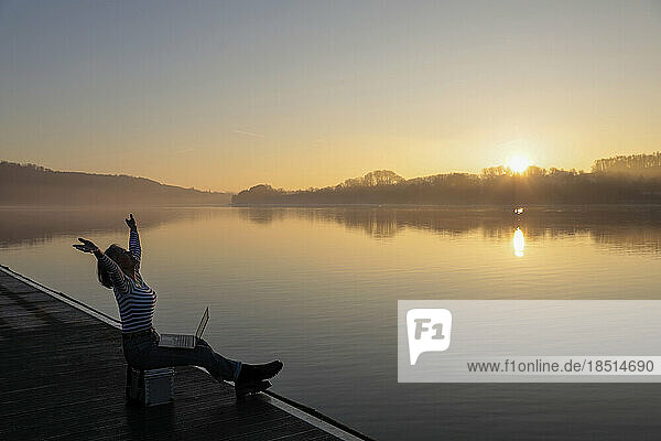 Freelancer with hands raised sitting with laptop by lake in morning