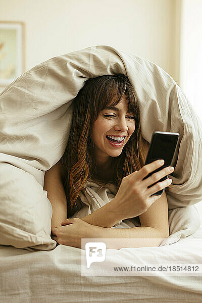 Happy woman using smart phone wrapped in blanket at home