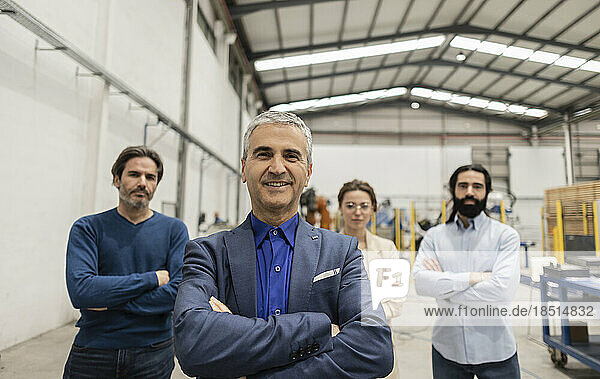 Smiling businessman standing with arms crossed in front of colleagues at factory