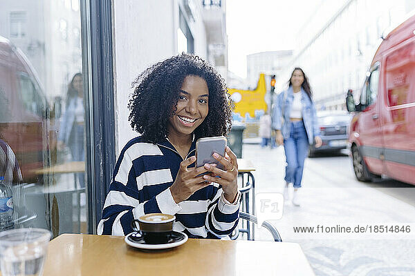 Happy young woman with smart phone sitting at sidewalk cafe