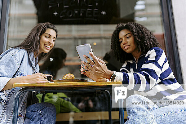 Happy young woman with friend using smart phone at sidewalk cafe