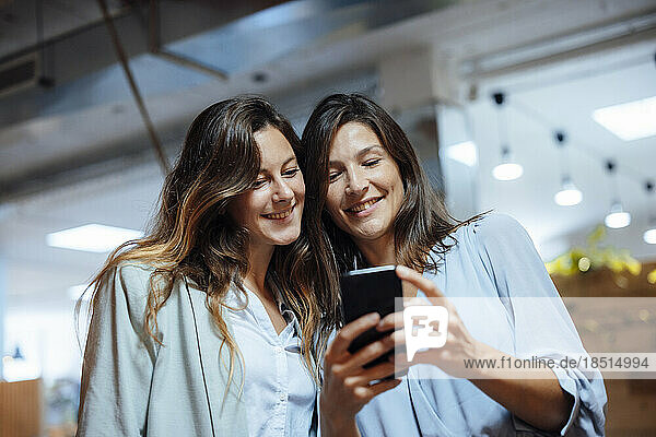 Happy businesswoman sharing smart phone with colleague in office