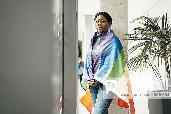 Young businesswoman wrapped in rainbow flag standing at office