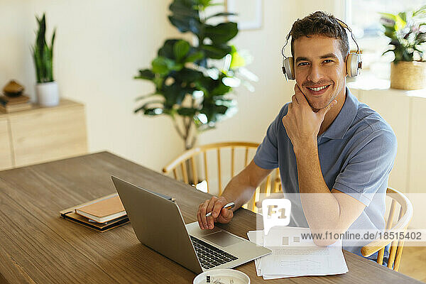 Happy freelancer wearing headphones sitting with laptop at table in home office