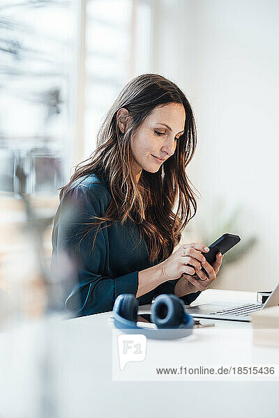 Lächelnde junge Geschäftsfrau  die im Heimbüro ihr Smartphone benutzt