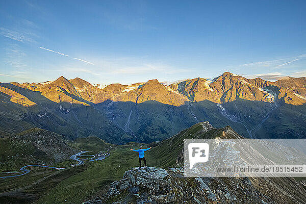 Austria  Salzburger Land  Grossglockner Road seen from summit of Edelweissspitze mountain at dawn with tourist standing with raised arms in foreground