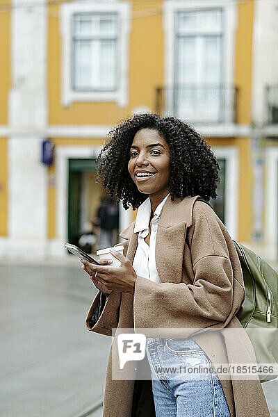 Happy young woman with smart phone and coffee cup standing in front of building