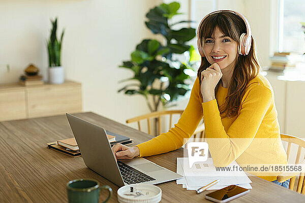 Happy woman wearing headphones sitting with laptop at table in home office