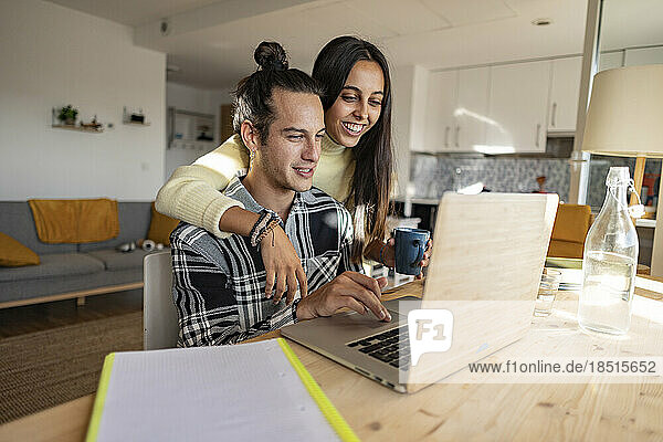 Happy girlfriend with arm around boyfriend using laptop at home office