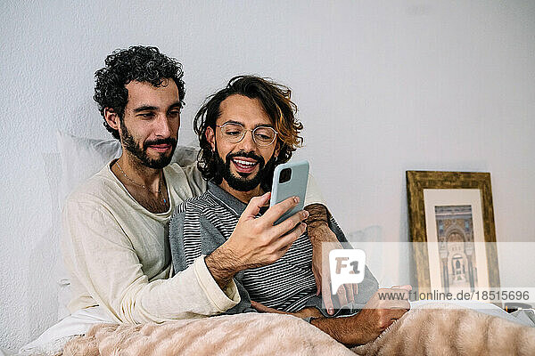 Man sharing smart phone with boyfriend sitting on bed at home