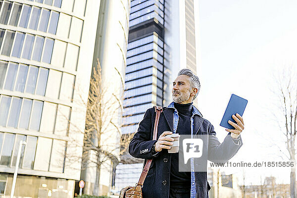 Mature businessman with tablet PC and coffee cup standing in front of buildings