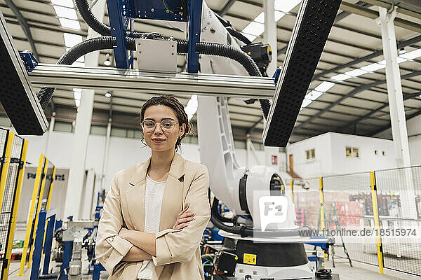 Young engineer standing with arms crossed in front of robotic arm