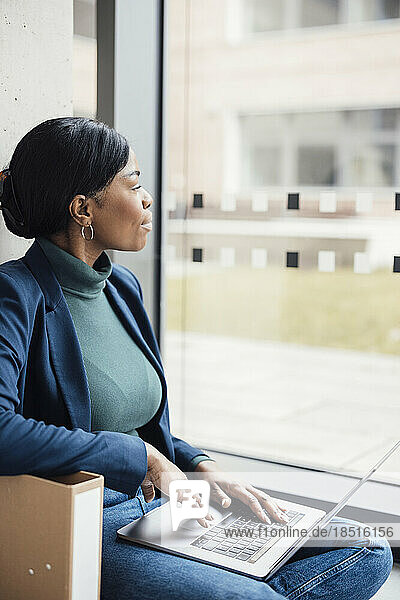 Businesswoman looking out of window sitting with laptop at office