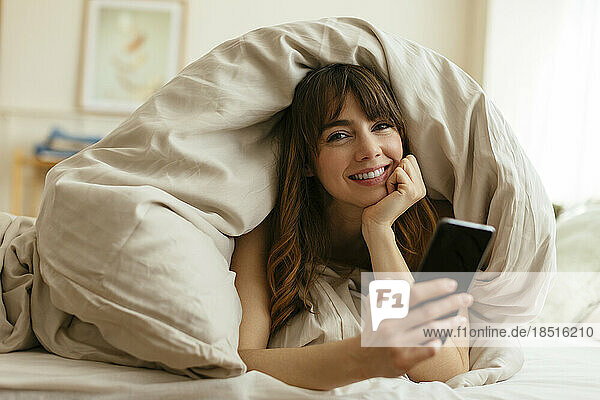 Happy woman with mobile phone wrapped in blanket at home