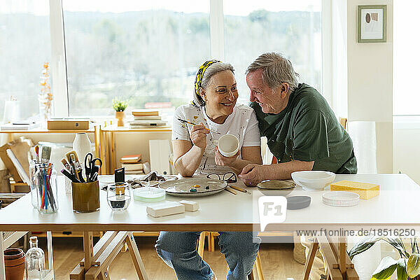 Happy senior man with woman painting ceramic cup in workshop