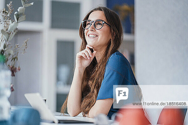 Lächelnde junge Geschäftsfrau mit der Hand am Kinn sitzt am Schreibtisch