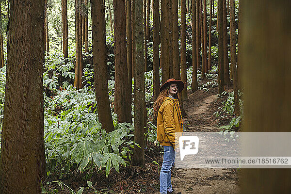 Rothaarige Frau mit Hut steht inmitten hoher Bäume im Wald