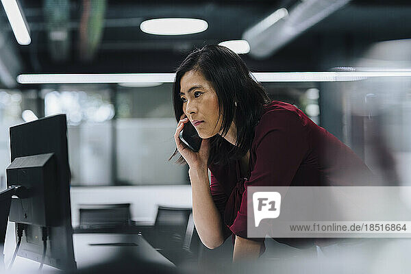 Businesswoman talking on smart phone leaning on desk in office