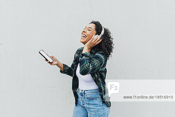 Cheerful woman wearing wireless headphones standing with smart phone