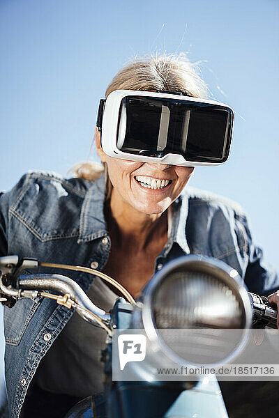Glückliche Frau mit VR-Brille  die unter blauem Himmel Fahrrad fährt
