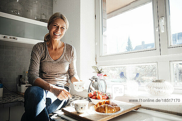 Happy woman having breakfast sitting on kitchen counter near window