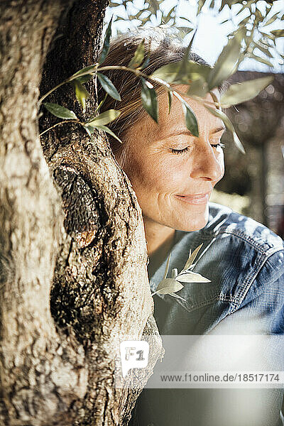 Smiling mature woman with eyes closed hugging tree on sunny day