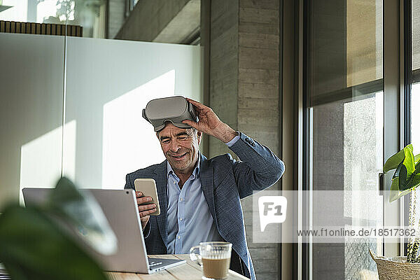 Mature businessman with VR glasses using smart phone in office