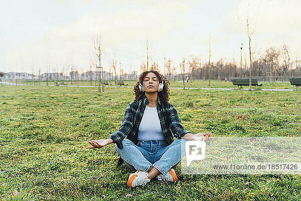 Young woman wearing wireless headphones meditating in park