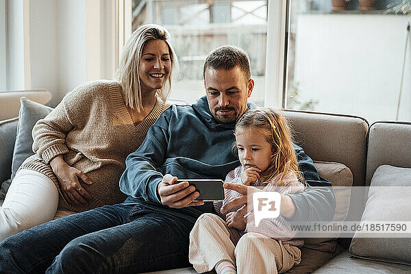 Parents sharing smart phone with daughter sitting on sofa at home